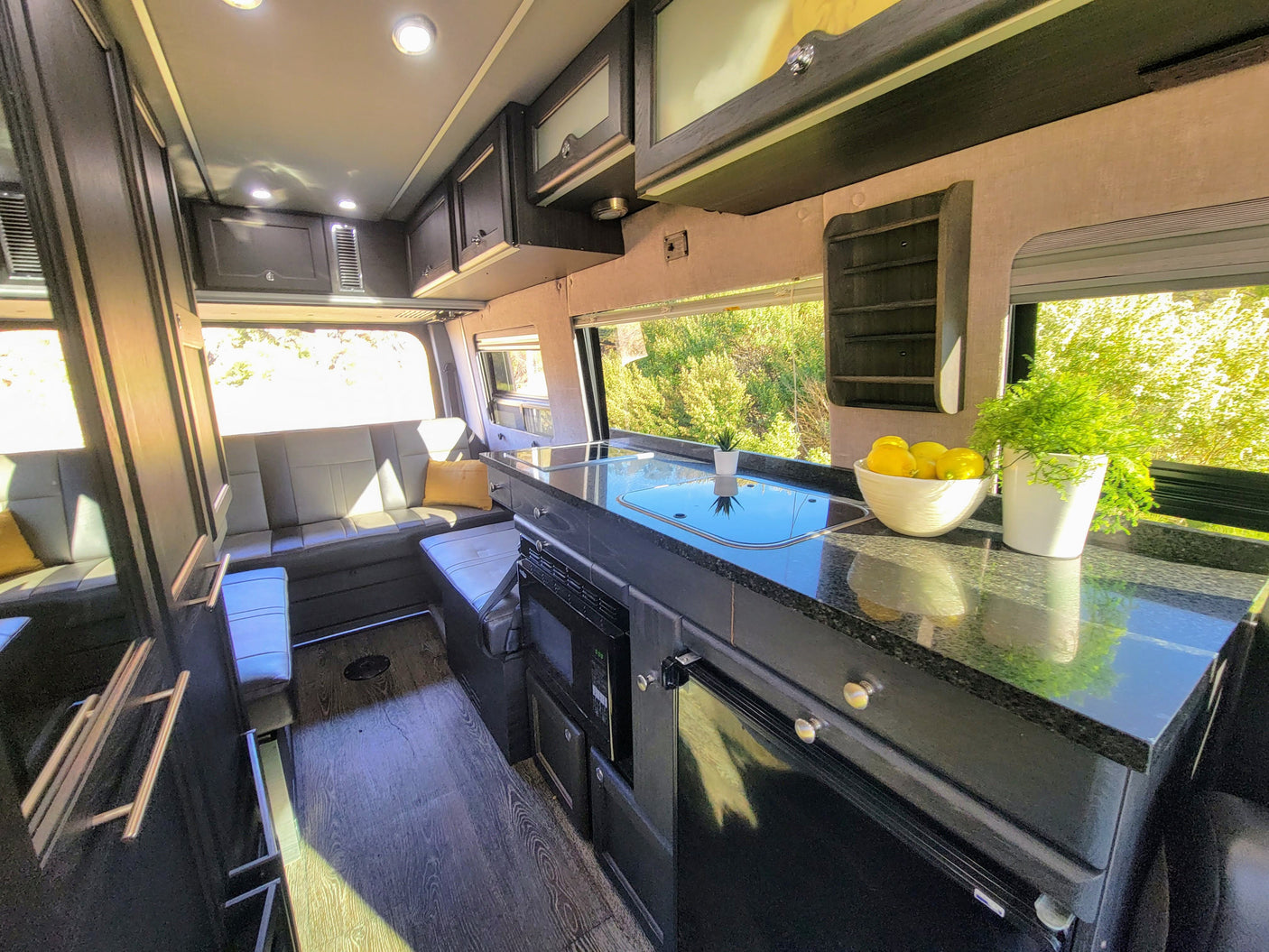 Spacious RV rental interior: kitchen with cooktop, sink, microwave; comfy bench seating, overhead storage, nature outside.