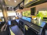 Spacious RV rental interior: kitchen with cooktop, sink, microwave; comfy bench seating, overhead storage, nature outside.