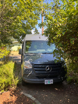 A gray Mercedes-Benz Sprinter van, likely an RV rental, is parked on a dirt driveway amidst green foliage under a clear sky.