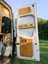 Rear of an RV rental van shows a water heater, propane tank, and plumbing setup. The open door reveals custom wood paneling.