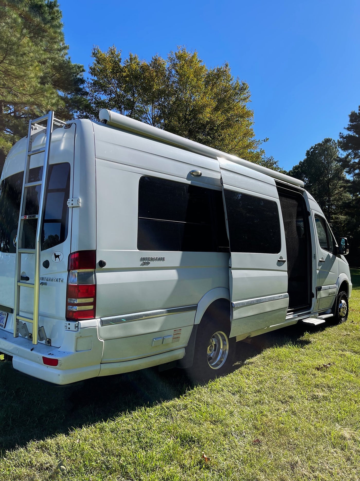 White RV rental parked on green grass under a clear blue sky, with its side door open and a ladder visible on the rear. — photo 3