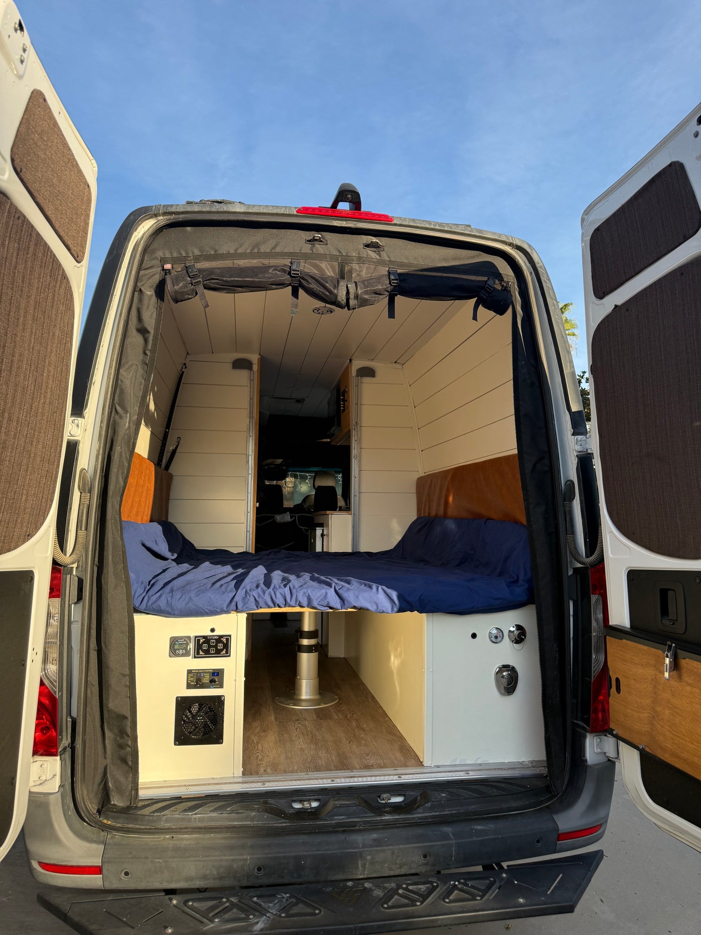 Step inside this cozy RV rental camper van! Features include a custom bed, control panel, and open rear doors.