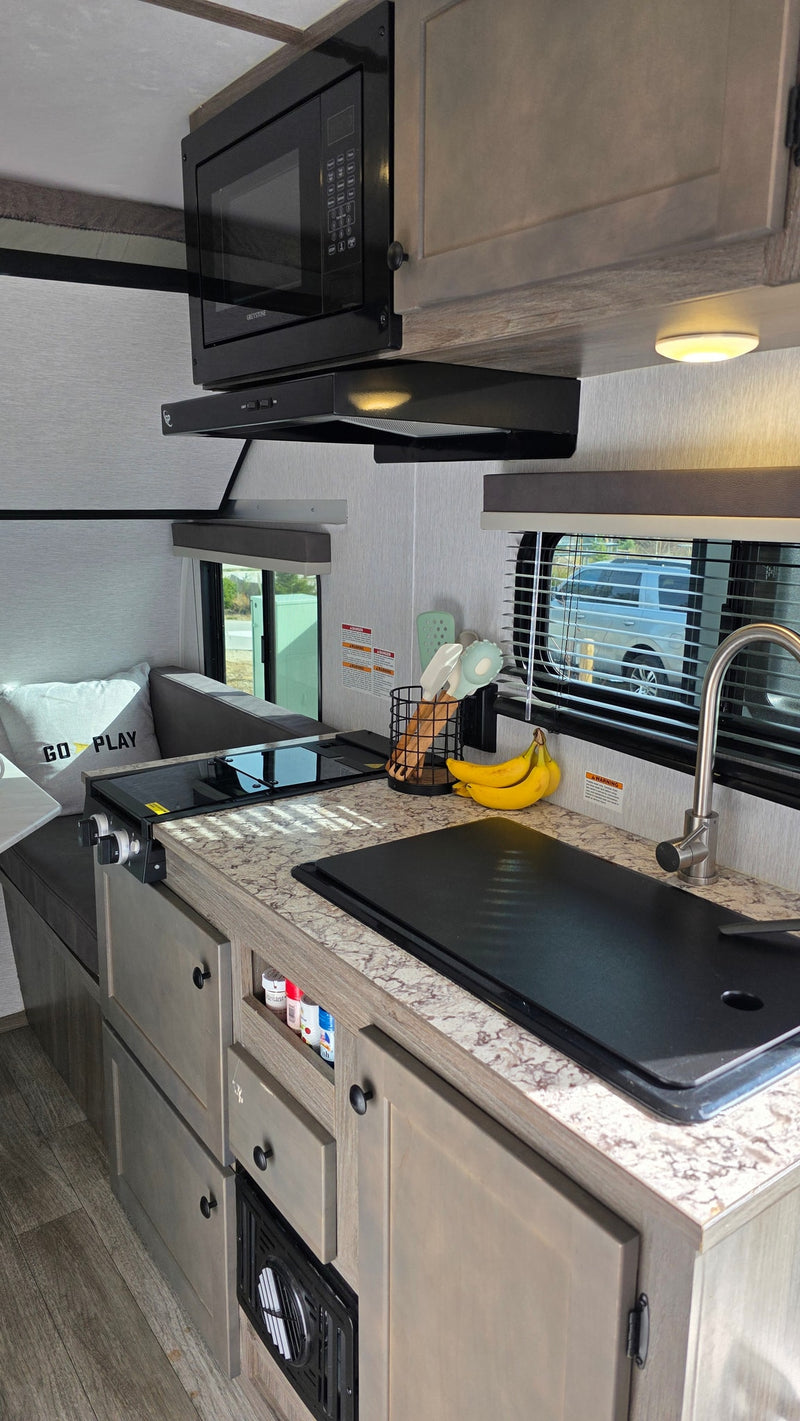 Explore this rv rental's compact kitchen, featuring a microwave, cooktop, covered sink, and wood-toned cabinets. — photo 3