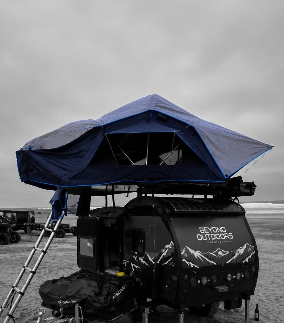  with a Jeep and ladder on sandy terrain.|Blue rooftop tent on an RV rental teardrop trailer set up for beach camping. A ladder extends to the sand.|This RV rental teardrop camper with its deployed rooftop tent is perfect for desert camping under a bright blue sky.|Beyond Outdoors RV rental setup: a rooftop tent — photo 3