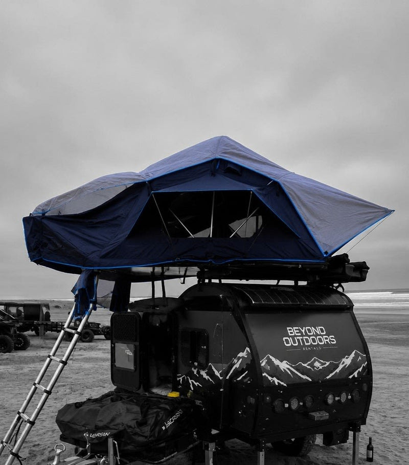  with a Jeep and ladder on sandy terrain.|Blue rooftop tent on an RV rental teardrop trailer set up for beach camping. A ladder extends to the sand.|This RV rental teardrop camper with its deployed rooftop tent is perfect for desert camping under a bright blue sky.|Beyond Outdoors RV rental setup: a rooftop tent — photo 3