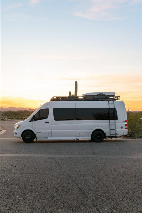 A white RV rental, a custom Sprinter van with roof rack & ladder, parked on a desert road at sunset.