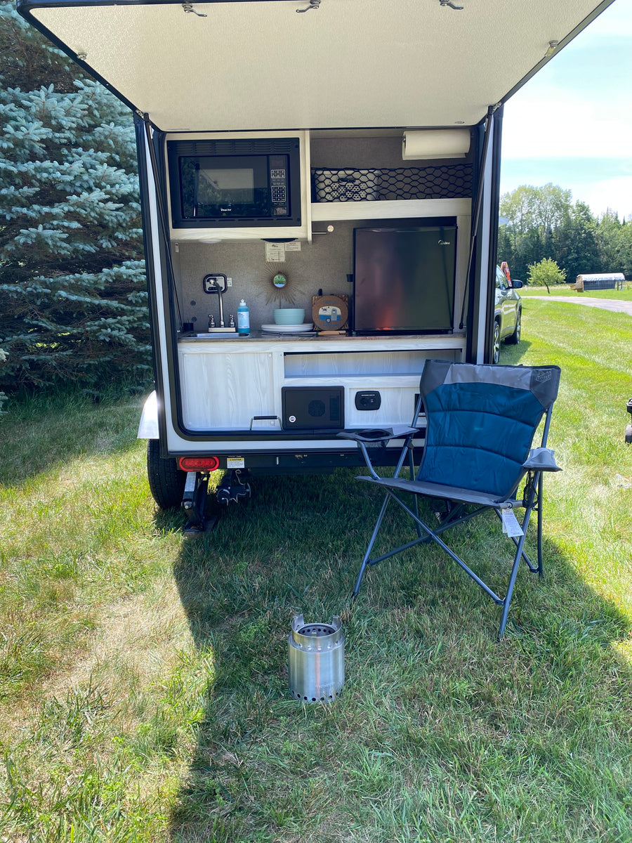 Compact RV rental with rear kitchen (microwave, fridge, sink) open for outdoor use. Camping chair & Solo Stove nearby.