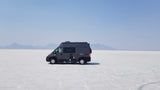 RV rental freedom: Dark grey camper van on vast white Bonneville Salt Flats, mountains under hazy sky.