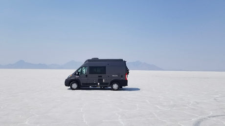 RV rental freedom: Dark grey camper van on vast white Bonneville Salt Flats, mountains under hazy sky.