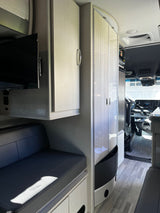 Modern rv rental interior: light wood-look cabinets, dark bench seating, TV, and the Mercedes-Benz cockpit visible.