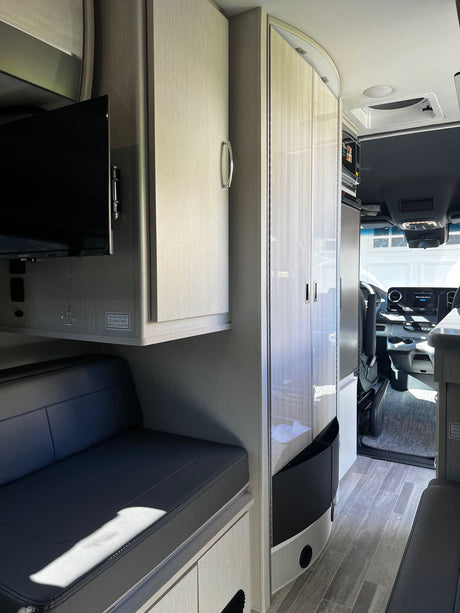Modern rv rental interior: light wood-look cabinets, dark bench seating, TV, and the Mercedes-Benz cockpit visible.