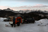 Scenic mountain RV rental trip: A person cooks at a teardrop trailer, parked with cars amidst snowy peaks.