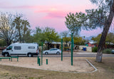 White RV rental parked on a residential street next to a park with exercise equipment under a pink sky.