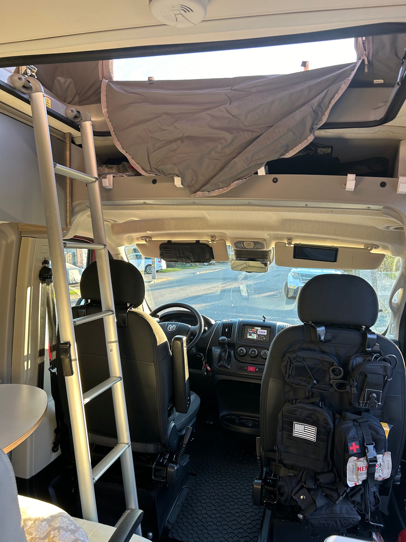 RV rental interior: front cabin with driver's seat, passenger seat with tactical gear, ladder to overhead bunk, and upper storage area.
