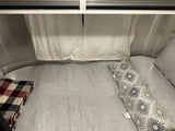 Cozy RV rental bed features light gray linens, a red plaid blanket, and patterned pillows. Perfect for a comfortable travel trailer trip.