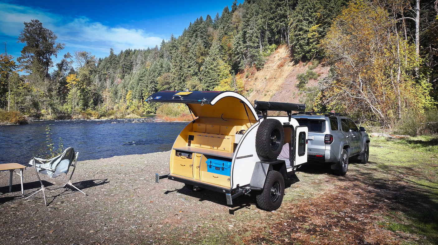 Overlanding setup: An RV rental teardrop camper and SUV by a scenic river, perfect for camping.