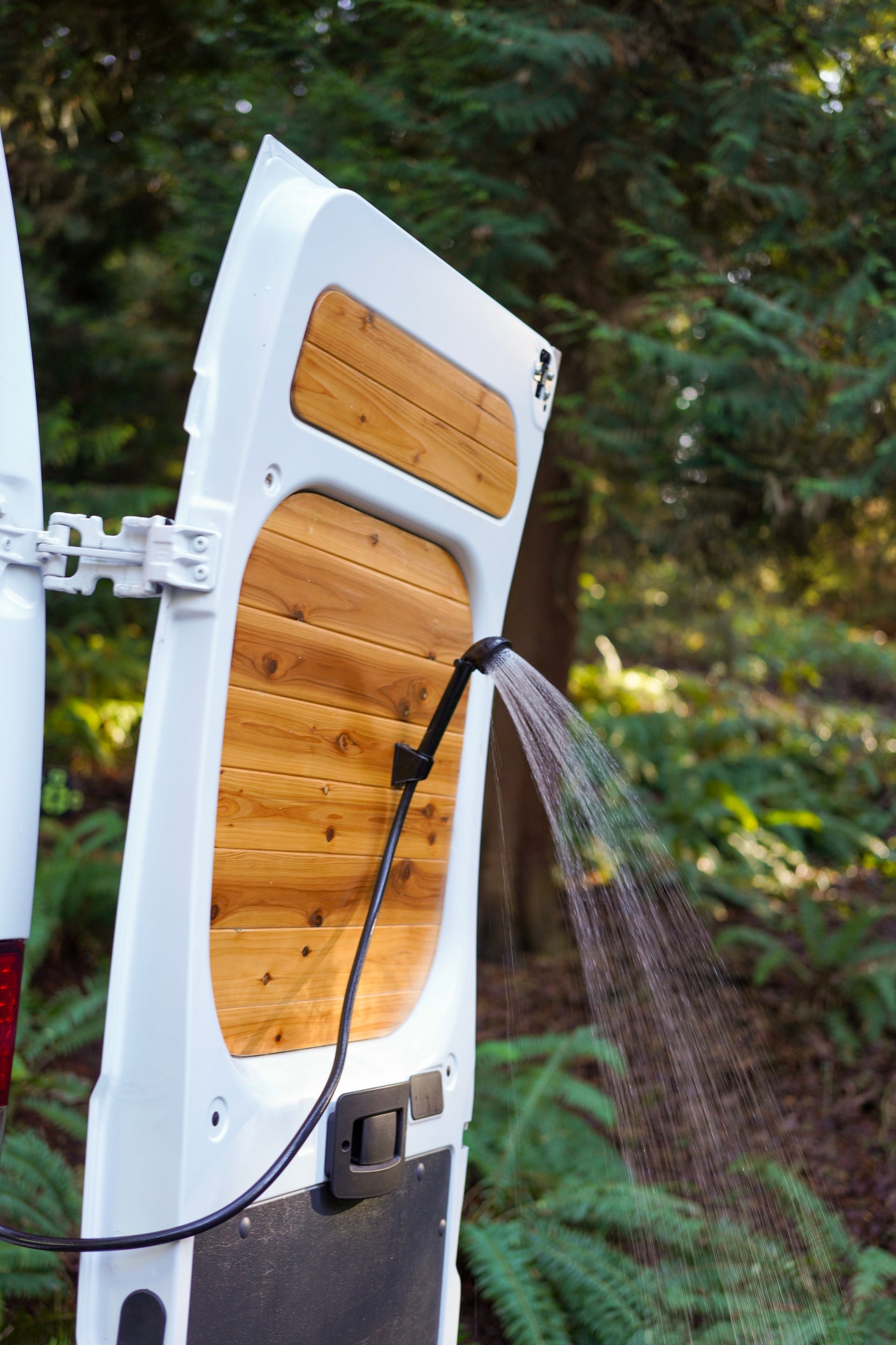 Outdoor shower spraying water from an RV rental's rear door with wood paneling, set against a blurred forest. Perfect for outdoor adventure.