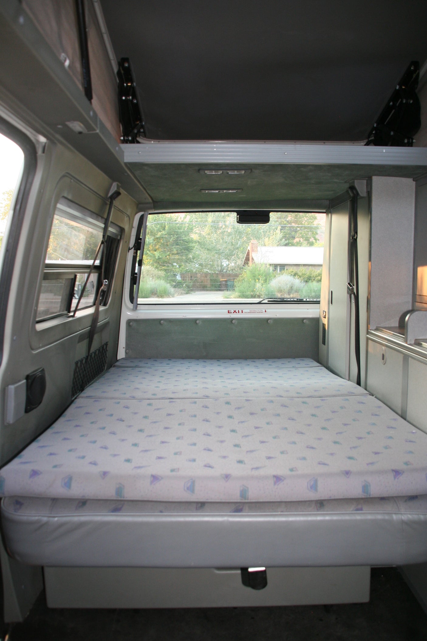 View inside an RV rental: A sleeping area with a patterned mattress on the right, a large window looking outdoors, and the vehicle's front cabin visible.