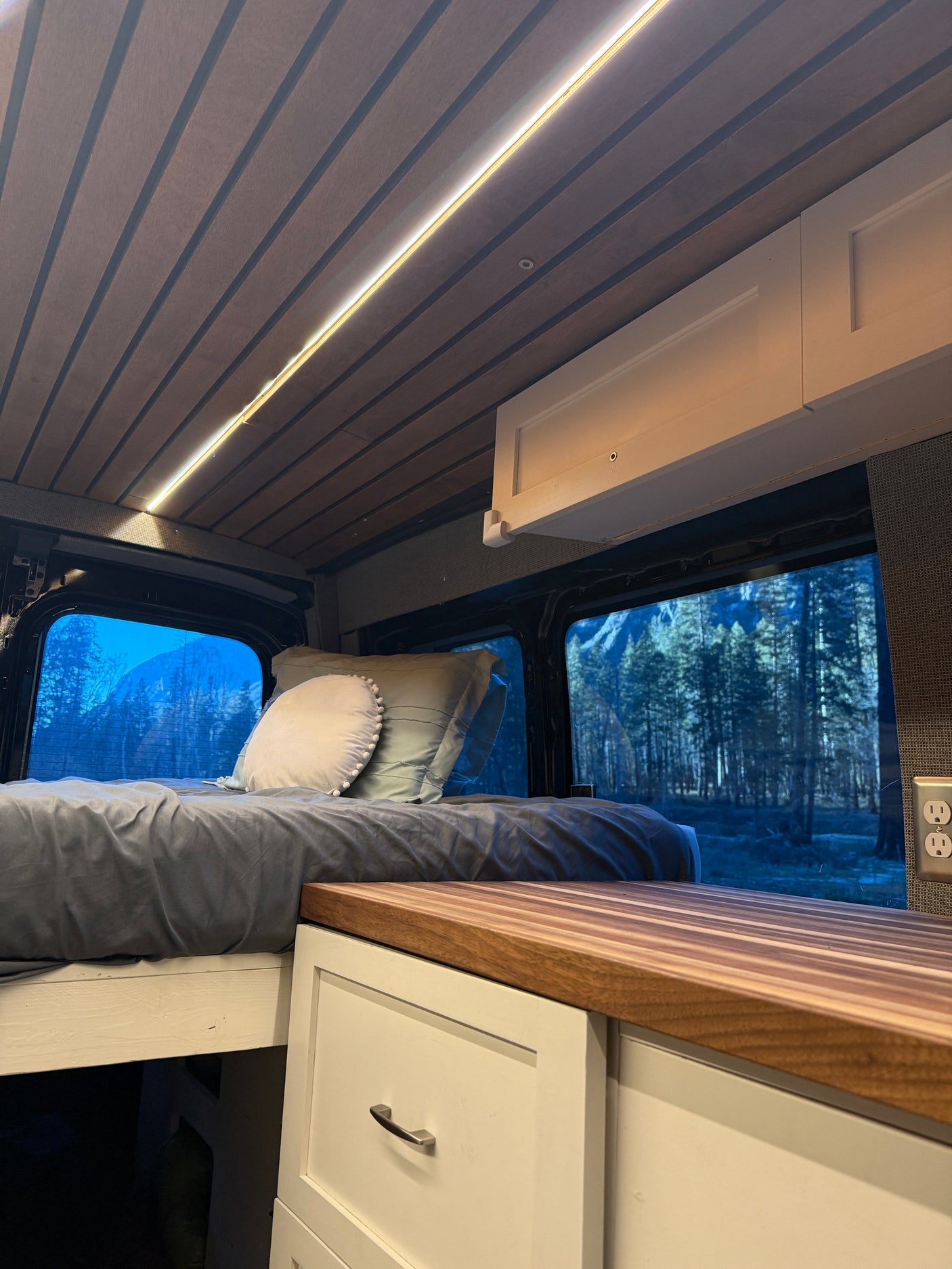 Cozy RV rental interior: bed, warm LED ceiling light, and a window framing a tranquil forest and mountains.