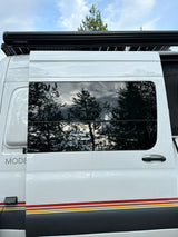 Side exterior of a white rv rental camper van, showing Dometic awning, reflective windows with trees, and colored stripes.