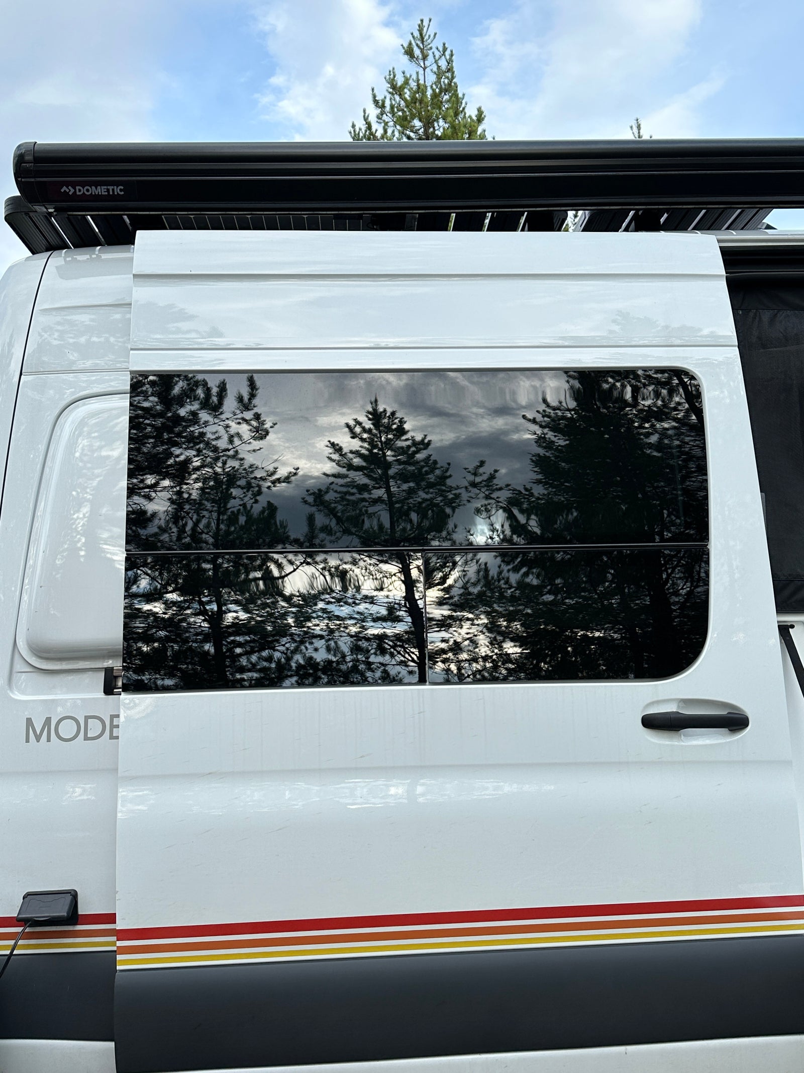 Side exterior of a white rv rental camper van, showing Dometic awning, reflective windows with trees, and colored stripes.