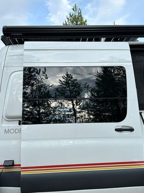 Side exterior of a white rv rental camper van, showing Dometic awning, reflective windows with trees, and colored stripes.