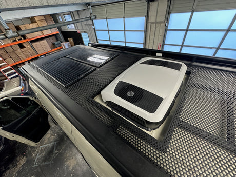 RV rental van's roof with solar panels, rooftop AC unit, and perforated platform, parked in a workshop setting.