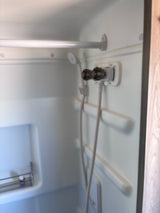 Compact white RV rental shower interior with a hose-style showerhead, water controls, and a small shelf.