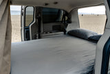 Interior view of an RV rental showing a cozy bed inside, with an open door revealing a sandy beach and ocean outside. Perfect for van life.