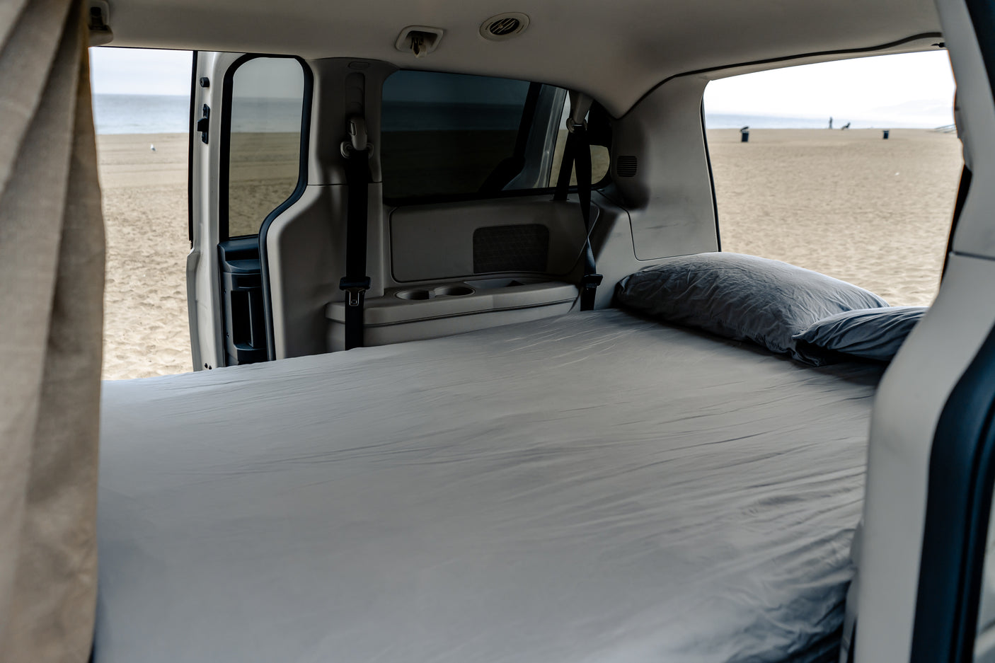 Inside this RV rental, a bed with grey sheets and pillows faces an open door, revealing a sandy beach and ocean view.