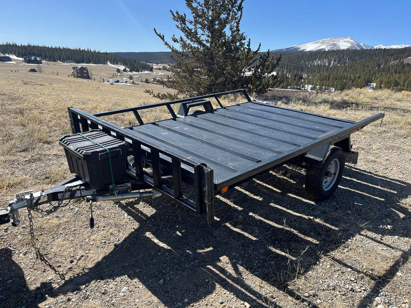 A black utility trailer, ideal for your RV rental, is parked on a dirt hill with scenic mountains and cabins in the distance. — photo 2