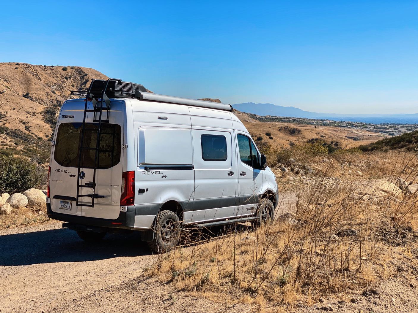 White Winnebago Revel camper van, an ideal RV rental, parked on a dirt path in dry, hilly terrain under a clear sky.
