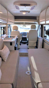 Bright RV rental interior showing a compact kitchen, lounge seating/bed area, and the driver's cabin beyond.