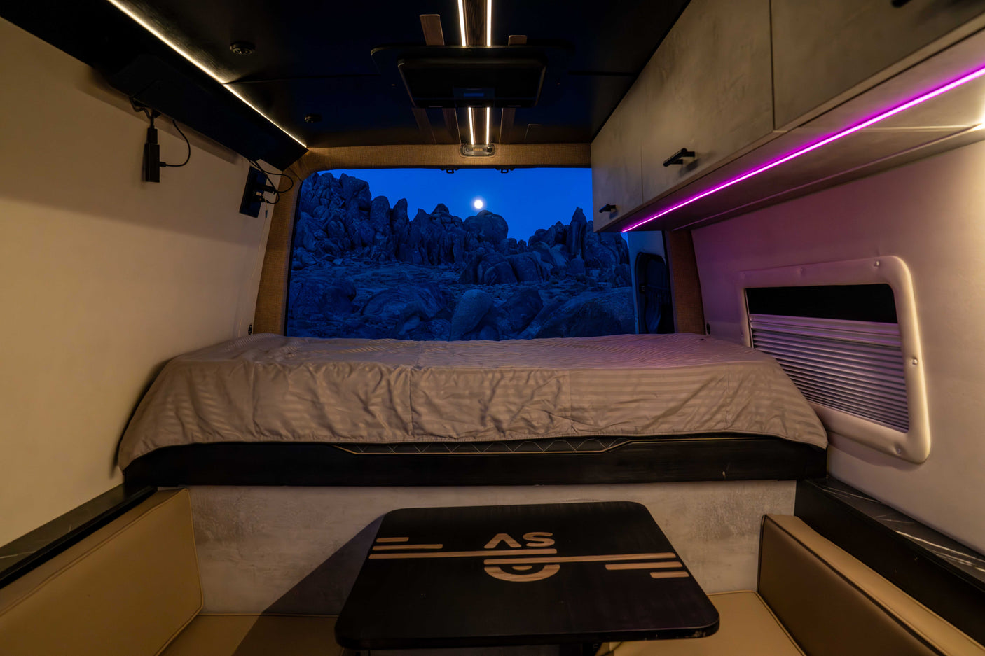 Step into this modern **rv rental**'s cozy interior, featuring a comfortable bed and a window framing a moonlit rocky landscape.