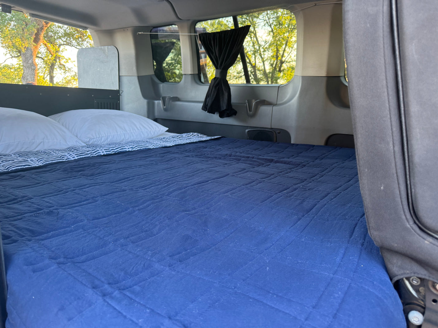 Inside an RV rental: a comfy bed with blue quilted bedding and white pillows. Windows with curtains offer tree views.