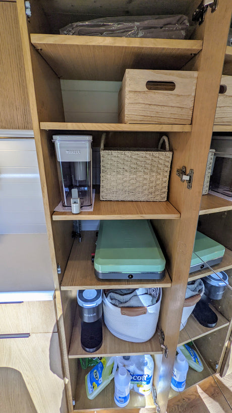 Organized RV rental cabinet displaying kitchen essentials: Brita, Keurig, portable stove, baskets, and cleaning supplies.