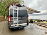 A grey Winnebago Travato rv rental parked by a scenic waterfront. Awning extended with lights and a camping chair.
