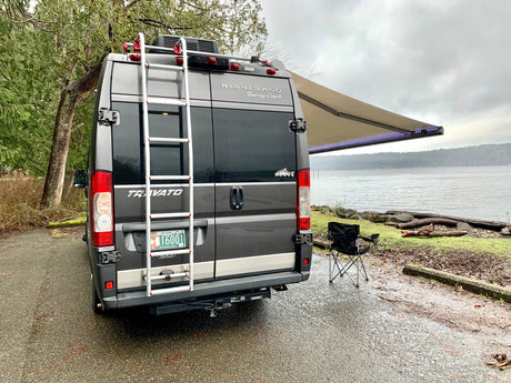 A grey Winnebago Travato rv rental parked by a scenic waterfront. Awning extended with lights and a camping chair.