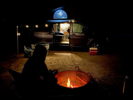 Night camping with an RV rental: a person enjoys a campfire, beside a lit camper van with a pop-top tent & string lights.
