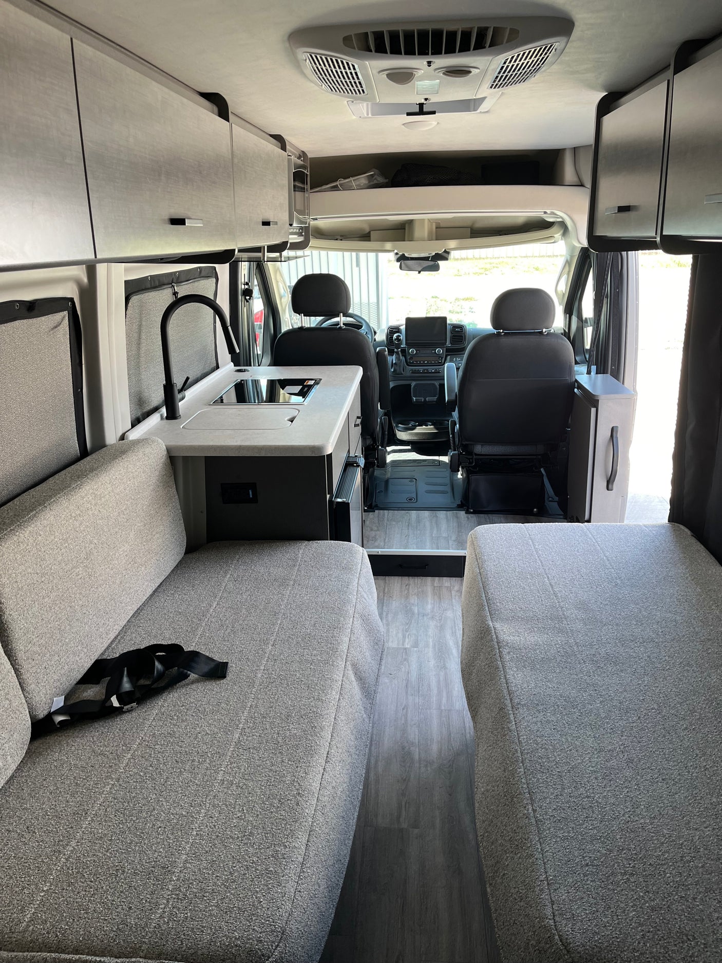 Modern rv rental interior with kitchenette, gray fabric seating, overhead cabinets, and front cab view. Sleek, organized space.