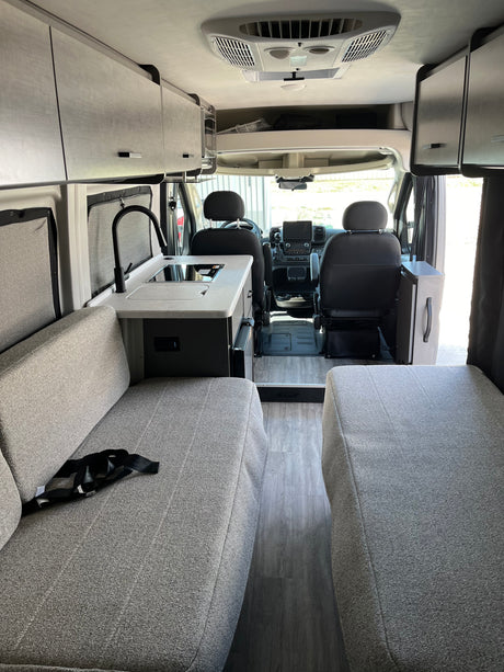 Modern rv rental interior with kitchenette, gray fabric seating, overhead cabinets, and front cab view. Sleek, organized space.
