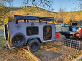 A gray Texas Adventure Rentals RV rental (teardrop camper) at a sunny campsite with a red truck, solar panel, and fall foliage.