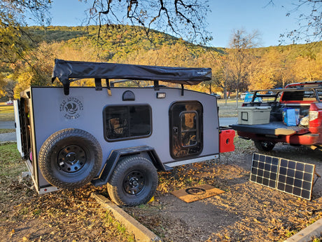 A gray Texas Adventure Rentals RV rental (teardrop camper) at a sunny campsite with a red truck, solar panel, and fall foliage.