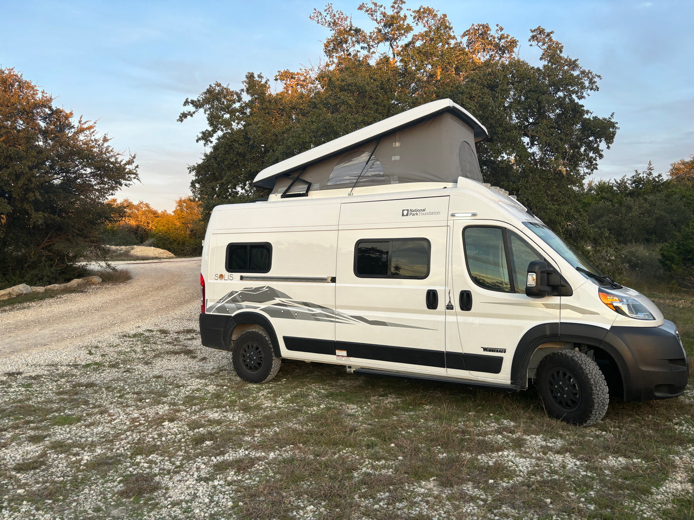 This white Winnebago Solis camper van with pop-top roof is parked on a dirt path for an RV rental outdoor adventure.