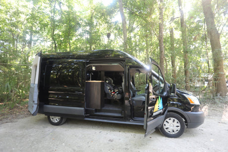 Black camper van for RV rental parked outdoors with doors open, revealing a sink and car seat inside.