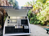 Outdoor kitchen setup of an RV rental at a sunny campsite, featuring a sink, storage, and portable fridge for convenient travel.