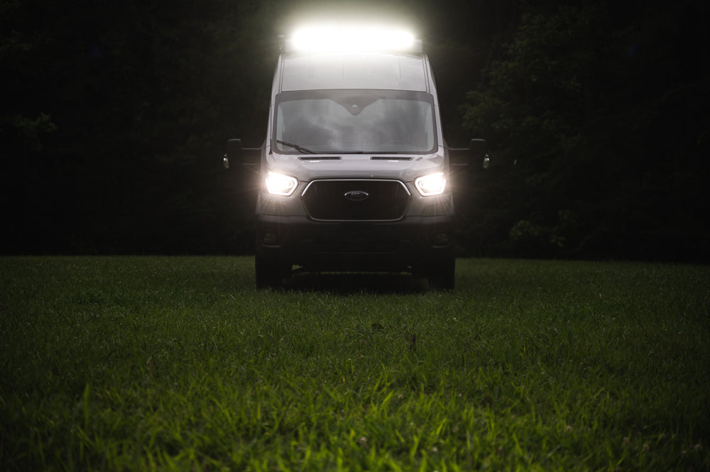 Ford Transit van, an RV rental, brightly lit by its headlights and roof light in a dark grassy field.