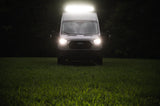 Ford Transit van, an RV rental, brightly lit by its headlights and roof light in a dark grassy field.