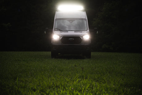 Ford Transit van, an RV rental, brightly lit by its headlights and roof light in a dark grassy field.