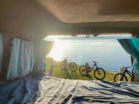 Serene RV rental view: cozy interior bed, ocean, and mountain bikes on grass. Ideal van life adventure.